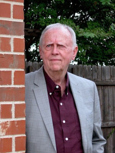 Photo of Caleb Pirtle, leaning against a red brick wall, dressed in a grey suit jacket with a maroon button up shirt beneath, looking off slightly to his right.